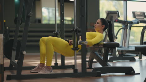 A-young-woman-in-a-yellow-tracksuit-lifts-her-body-with-a-barbell-to-train-the-back-of-her-thigh-and-buttocks.-Weight-training-in-the-smith-simulator.-Lifting-the-waist-and-body-with-weight-in-the-gym